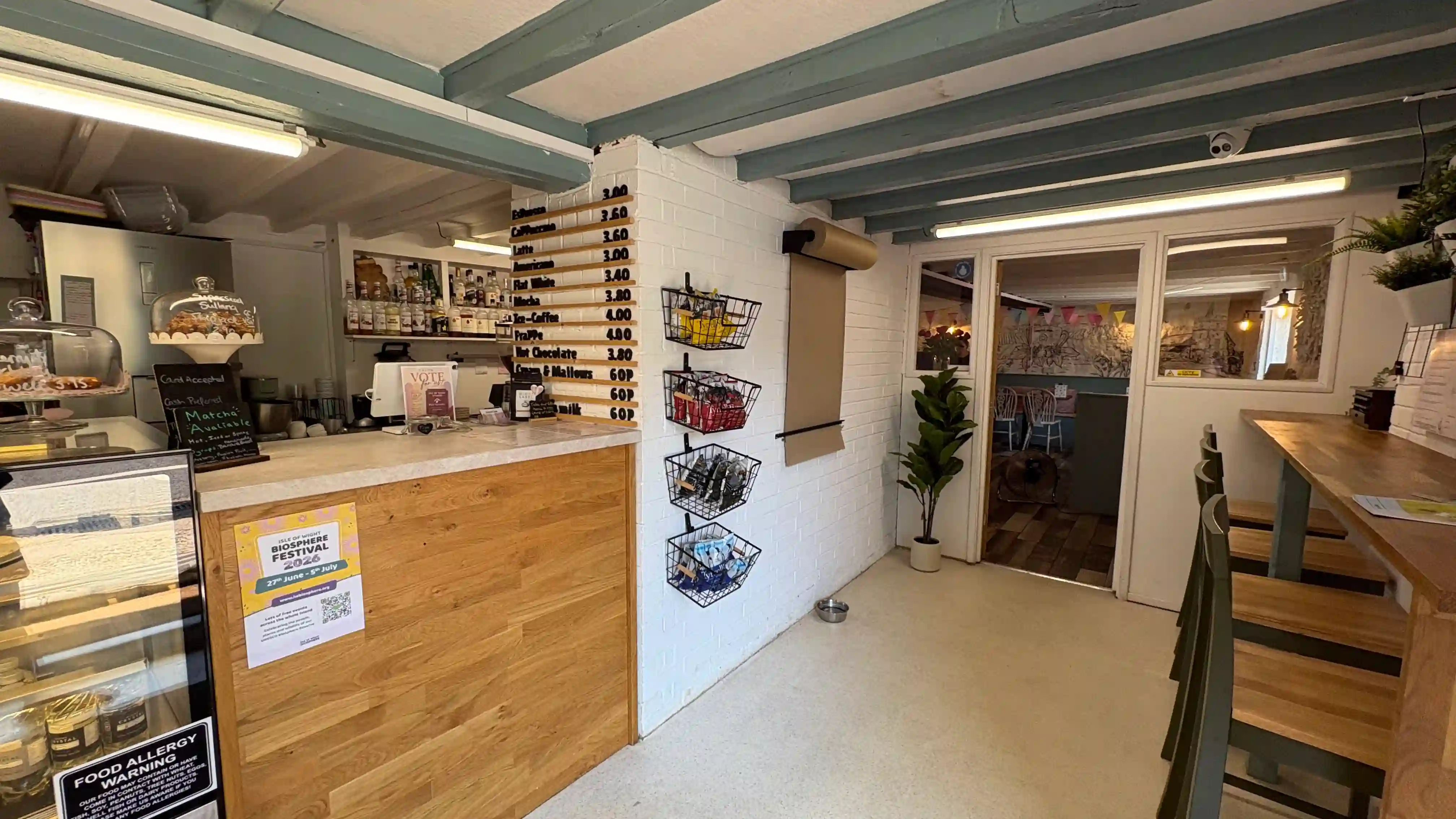 The slatted oak counter with cake domes, cake cabinet and wooden slatted drinks board, with Katy Rose's black-ink mural visible through the doorway to the back dining room